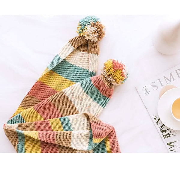 Colorful striped beanie with pom-poms on a white surface with a magazine and cup.
