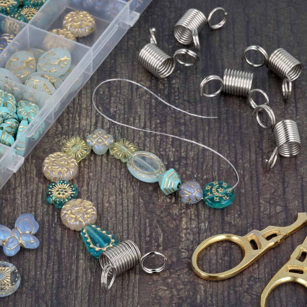 Jewelry making supplies arranged on a dark wood surface, including silver coil end pieces, clear wire, faceted beads in turquoise, cream, and gold, round embellishments, a clear compartment box of beads, and gold-toned scissors.