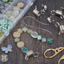 Beaded wire jewelry components laid out on a dark wood surface, including turquoise and gold patterned beads, silver coil wraps, clear elastic cord, metal rings, and a clear plastic compartment box of beads.