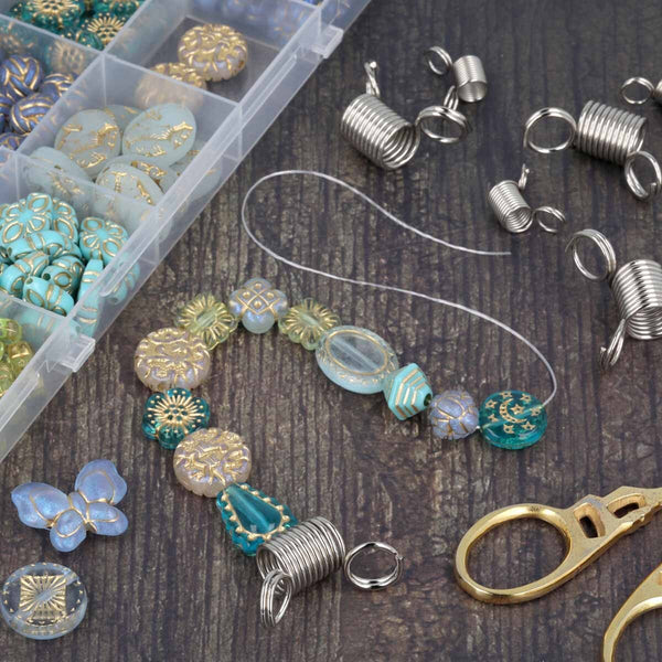Beaded wire jewelry components laid out on a dark wood surface, including turquoise and gold patterned beads, silver coil wraps, clear elastic cord, metal rings, and a clear plastic compartment box of beads.