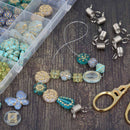 A flat-lay of craft beads and jewelry-making components on a dark wood surface, including turquoise and gold patterned beads, silver wire jump rings, a clear plastic bead organizer, and gold-handled scissors.