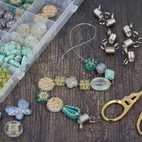 A flat-lay of craft beads and jewelry-making components on a dark wood surface, including turquoise and gold patterned beads, silver wire jump rings, a clear plastic bead organizer, and gold-handled scissors.