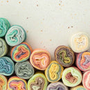 A collection of colorful variegated yarn balls arranged in a curved row along the bottom of a light beige speckled surface, leaving negative space above.