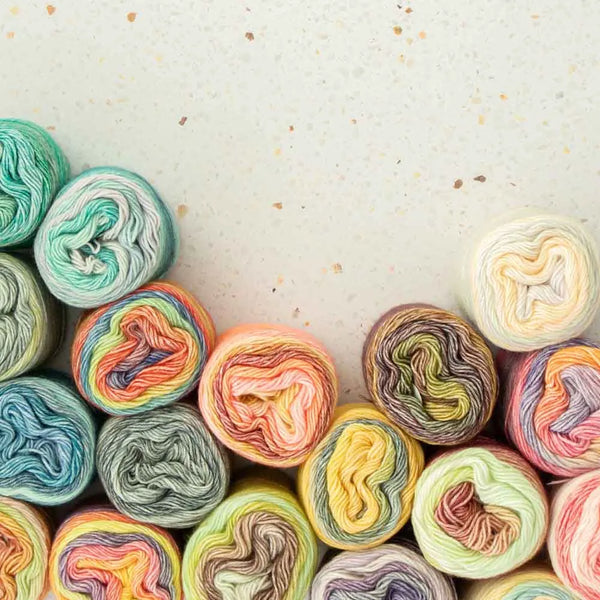 A collection of colorful variegated yarn balls arranged in a curved row along the bottom of a light beige speckled surface, leaving negative space above.