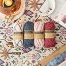 Four skeins of Scheepjes Stone Washed Merino wool yarn in cream, navy, pink, and burgundy, arranged on a patterned textile with a candle, scissors, and an open book nearby.