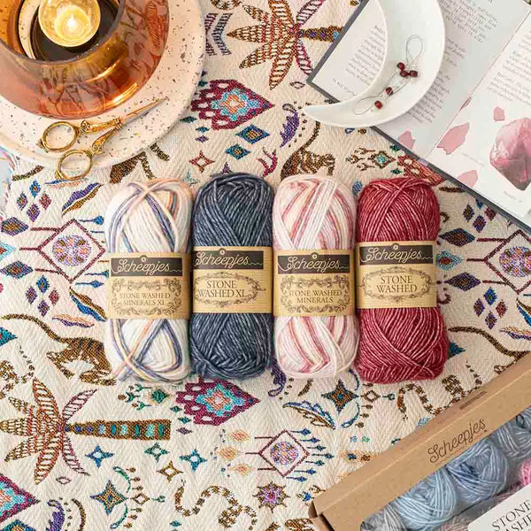 Four skeins of Scheepjes Stone Washed Merino wool yarn in cream, navy, pink, and burgundy, arranged on a patterned textile with a candle, scissors, and an open book nearby.