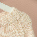 Close-up of a cream-colored knitted sweater with a ribbed and cable-knit pattern, shown on a white hanger against a soft pink background.
