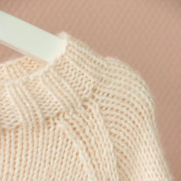 Close-up of a cream-colored knitted sweater with a ribbed and cable-knit pattern, shown on a white hanger against a soft pink background.