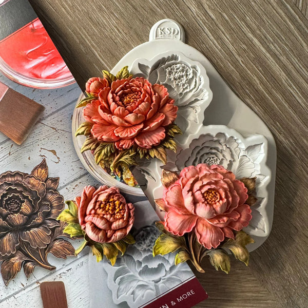 A white silicone mold tray with floral cavities, shown alongside three 3D-printed peony flower models in coral, pink, and peach tones with green and yellow leaves. The scene is styled on a wood-grain tabletop with craft-related props, including a red palette and wooden blocks.