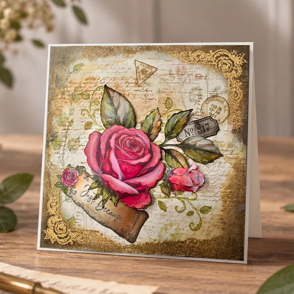 A square greeting card with a vintage-style floral design, featuring a large pink rose, smaller rosebud, green leaves, ornate gold borders, and script text on a warm beige background, photographed standing on a wooden surface.