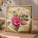 A square greeting card with a vintage-style floral design, featuring a large pink rose, smaller rosebud, green leaves, ornate gold borders, and script text on a warm beige background, photographed standing on a wooden surface.