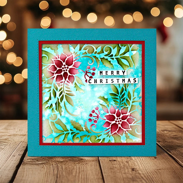 A square handmade Christmas greeting card with a turquoise border and a layered floral design, displayed on a wooden surface with warm bokeh lights in the background. The card features red poinsettias, green leaves, and a white banner that reads 'MERRY CHRISTMAS'.