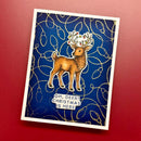 A square handmade greeting card featuring a brown deer with antlers decorated with Christmas lights, set against a dark blue background with gold abstract swirls and the text 'OH, DEER! CHRISTMAS IS HERE'. The card is photographed at an angle on a solid red surface.