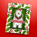 A square Christmas card featuring a white polar bear wearing a red Santa hat and green wreath with a red bow, centered on a dark green background. The card is framed by a white border with green holly leaves and red berries, with a red banner reading “BEARY CHRISTMAS.