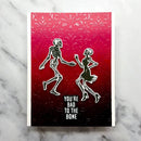 A rectangular greeting card with a red-to-black gradient design, featuring two dancing skeleton figures and the text 'YOU'RE BAD TO THE BONE'. The card is photographed flat on a light gray marble surface.