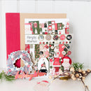 A Christmas-themed scrapbooking or card-making kit displayed on a white surface against a white paneled background. The kit includes a kraft paper sheet, a red binder or folder, decorative stickers, buttons, a wreath embellishment, and holiday-themed paper cutouts, including a Santa face and a red Christmas hat.