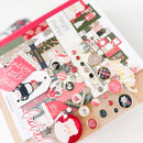 A flat-lay of a scrapbook or journaling kit featuring various Christmas-themed papers, stickers, buttons, and embellishments on a kraft card base.