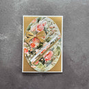 An oval handmade greeting card featuring a collage of pink tulips, green leaves, and a gold ribbon bow, set against a textured dark gray background.
