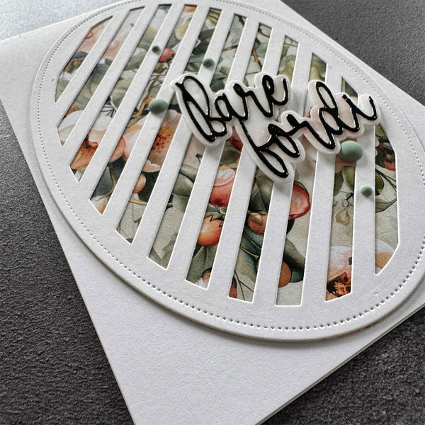 A close-up of a handmade greeting card featuring a white oval frame with vertical slats over a floral patterned insert, with a die-cut black script overlay reading 'bare feet' and small green bead accents, placed on a white backing card over a dark gray textured surface.