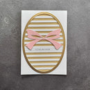 A handmade greeting card featuring a white background with horizontal gold and white stripes, an oval gold border, a pink paper bow, and the text 'TILLIKKE MED DAGAN' in black, photographed on a textured gray surface.