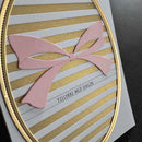 A close-up of a handmade greeting card featuring a pink paper bow on a background of white and glittery gold horizontal stripes, framed by an oval gold border. The text 'TILLYKÆ MED DAGEN' is printed in black.
