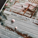 A close-up of a handmade Christmas greeting card or scrapbook paper featuring horizontal slatted wood textures, a watercolor winter scene with a ladder, pine branches, and the text 'MERRY CHRISTMAS TO YO'.