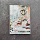 A rectangular Christmas greeting card featuring a snowy porch scene with a wreath, rocking chairs, and the text 'MERRY CHRISTMAS TO YOU AND YOURS', photographed against a dark textured background.
