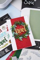 A handmade Christmas card featuring a red and white polka dot background with a green wreath and gold glittery 'Merry' lettering, styled on top of craft supplies including Sizzix and Tim Holtz papers.