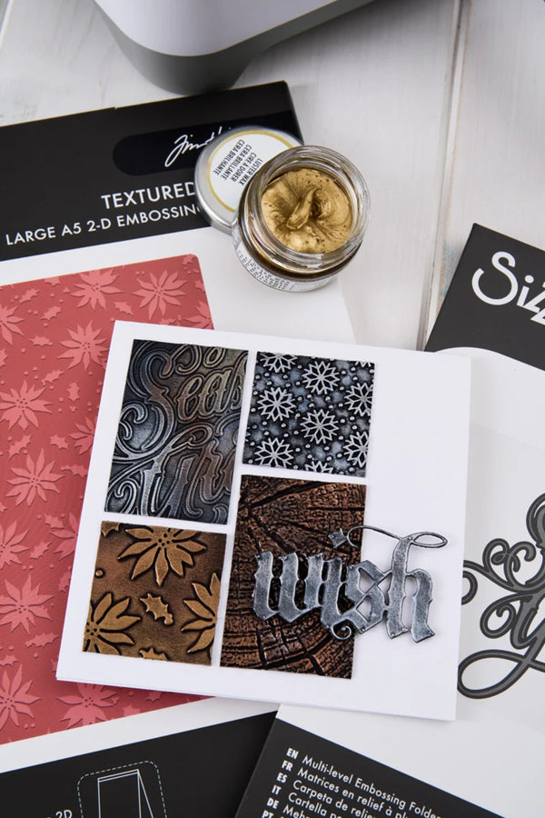 A styled flat-lay of a handmade greeting card featuring four embossed patterned squares in gray, black, and brown, with a silver metal 'wish' script overlay. The card is surrounded by crafting tools and papers, including a jar of textured gold embossing powder, a red patterned sheet, and black embossing folders.