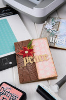 A handmade greeting card with a brown bark texture, a pink snowflake patterned panel, a red poinsettia flower, and a wooden 'peace' cutout, styled on a white surface with crafting tools and Tim Holtz paper packaging.