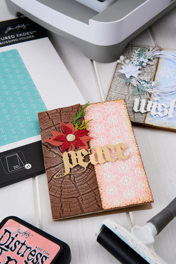 A handmade greeting card with a brown bark texture, a pink snowflake patterned panel, a red poinsettia flower, and a wooden 'peace' cutout, styled on a white surface with crafting tools and Tim Holtz paper packaging.