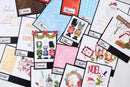 A flat-lay arrangement of multiple Christmas-themed card designs and scrapbooking papers on a white wooden background. The designs feature festive illustrations of Santa Claus, snowmen, nutcrackers, wreaths, and holiday greetings, with some cards showing black borders and others displaying colorful patterns.