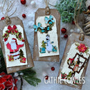Three handmade Christmas gift tags with festive layered paper cutouts, hanging from twine, arranged on a snowy white surface with holiday greenery and red ornaments. The text 'CATHIE COWLES' appears at the bottom right.