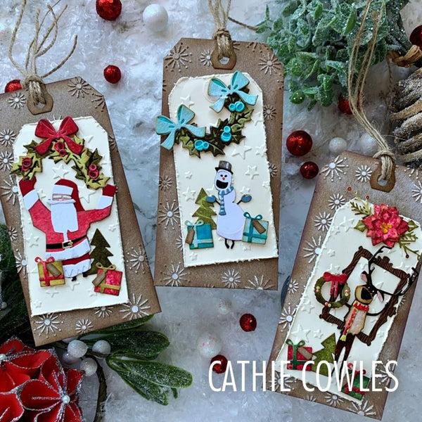 Three handmade Christmas gift tags with festive layered paper cutouts, hanging from twine, arranged on a snowy white surface with holiday greenery and red ornaments. The text 'CATHIE COWLES' appears at the bottom right.