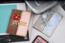 Two handmade greeting cards displayed on a white surface next to crafting tools, including an embosser, a stamping plate, and an ink pad. The left card features a brown tree bark texture, a pink patterned center, a red flower, and gold 'peace' lettering.