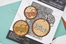A printed paper folder/package for a 3D embossing folder shown on a styled flat-lay with teal paper and a brown card. The package features three sample hanging round wood-grain tags with embossed words: 'now', 'wish', and 'peace' in red, gold, and silver tones.