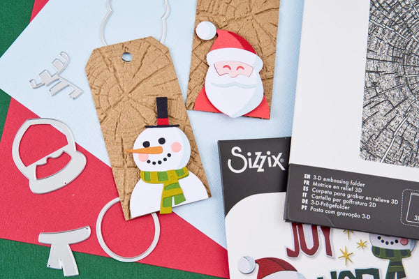 A styled flat-lay of Christmas-themed crafting supplies, including paper tags with a snowman and Santa design, metal die-cut shapes, and a package labeled 'Sizix' on a red, green, and white background.