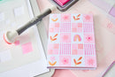 A pink and white patterned notepad with a patchwork design of flowers, leaves, and rainbows, styled on a desk with a pink ink pad and a gray marker.