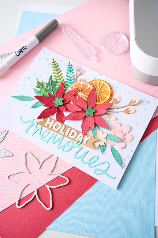 A handmade layered card with red poinsettias, green leaves, orange slices, and the words “Holiday Memories” in light blue script, styled on pastel pink and blue paper with craft tools nearby.
