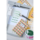 Crafting tools and materials including a squeegee, cutting mat, and decorative paper on a white background.