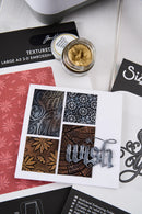 A styled flat-lay of a handmade greeting card featuring four embossed patterned squares in gray, black, and brown, with a silver metal 'wish' script overlay. The card is surrounded by crafting tools and papers, including a jar of textured gold embossing powder, a red patterned sheet, and black embossing folders.