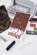 A printed card or art print with a dark ornate damask pattern in brown and orange above the word 'wicked' in a dripping red font, displayed on a white surface with a die-cutting machine, ink pad, and other craft supplies nearby.