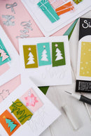 A styled flat-lay of handmade Christmas greeting cards and crafting tools on a white wooden surface. The central card features three vertical panels in olive green, teal, and dark green with white tree silhouettes and a star, above cursive 'Merry' text.