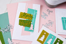 A styled flat-lay of handmade Christmas greeting cards and die-cut embellishments on a pink surface. The main card features layered pastel pink, mint green, and olive green stripes with metallic gold 'Merry Bright' script, a white bow, and a white Christmas tree.