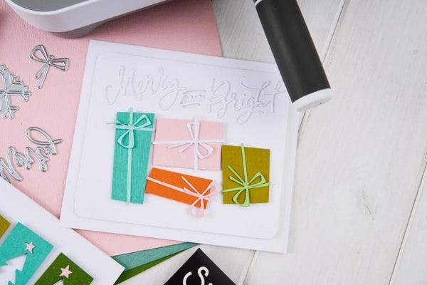 A flat-lay of a handmade greeting card featuring three colorful gift boxes with bows in teal, pink, and olive green, with a silver metal 'Merry & Bright' cutout on a white cardstock, surrounded by crafting tools and papers on a light wood surface.