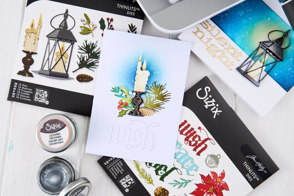 A styled flat-lay of crafting supplies on a white wood surface, including a central white card with a candle illustration and the word 'wish,' several ThinLustre Dies stamp cards, Sizzix heat transfer sheets, small Sizzix ink tins, and a silver laptop corner.
