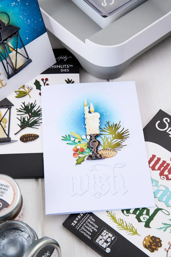 A handmade white greeting card featuring a watercolor illustration of two lit candles surrounded by pine branches, evergreen needles, and small red berries, with embossed script text reading 'wish' at the bottom. The card is styled on a white surface with craft supplies, stamps, and ink jars around it.