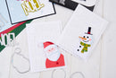 Two handmade greeting cards displayed on a white wooden surface, one featuring a layered Santa Claus design and the other a snowman design, with metal die-cutting and scoring tools nearby.