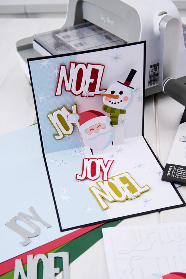 A handmade 3D pop-up Christmas greeting card displayed in a crafting workspace. The card features a Santa Claus and a snowman with layered text reading 'NOEL JOY NOEL' in red, white, and gold.