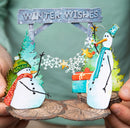 A handmade winter-themed paper cutout decoration featuring two snowmen, pine trees, a 'WINTER WISHES' sign, and wrapped gifts, held by hands against a soft-focus background.